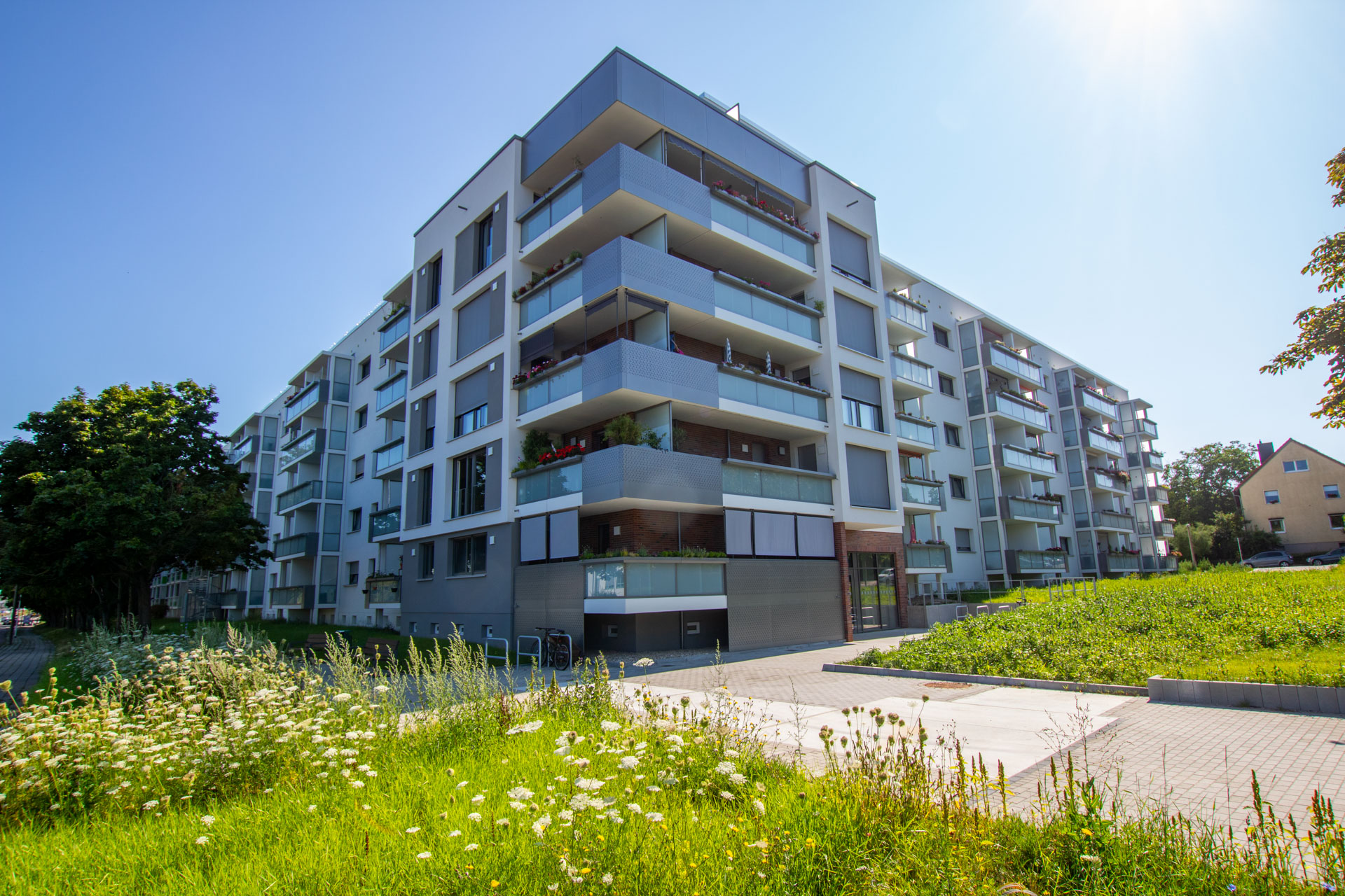 hellgrauer Wohnblock mit Balkonen im Neukirchener Weg bei Sonnenschein und blauem Himmel und einer Blühwiese